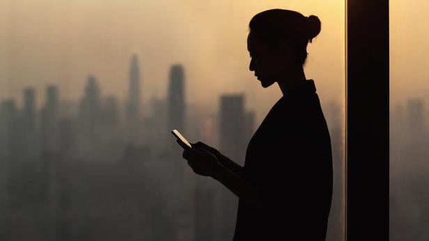 Silhouette of young woman using smartphone next to window with cityscape